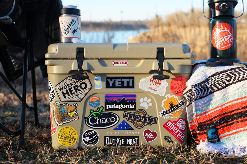 A beige cooler is covered in various colorful stickers, including brands like Yeti, Chaco, and Patagonia. It sits on grass by a lakeshore with a colorful blanket, a can, and a lamp nearby. | FintechZoom