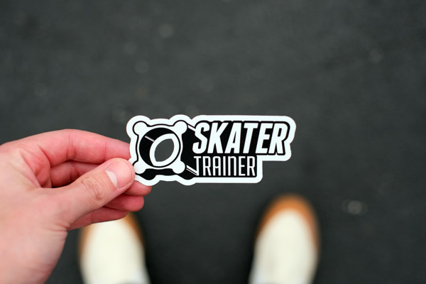 A hand holds a "Skater Trainer" sticker against a textured, dark asphalt background. The sticker is black and white with bold letters. White sneakers are partially visible at the bottom of the image. | FintechZoom