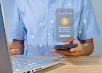 A person in a light blue shirt uses a smartphone at their desk with a laptop. A digital overlay displays "Online Banking" with a transfer button and a dollar sign icon, highlighting the convenience of White-Label Payment Gateways. | FintechZoom