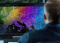 A person sits at a desk, looking at a computer monitor displaying a colorful, detailed map of Europe, with distinct color gradients indicating different regions or data zones. | FintechZoom