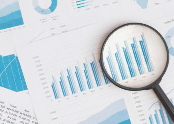 A magnifying glass placed over financial charts and graphs highlights blue bar graphs and data analysis on printed documents. Other charts, including pie and line graphs, are visible in the background. | FintechZoom