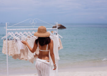 A woman in a sun hat and white crochet swimwear browses a rack of light-colored clothing on a sandy beach with calm blue ocean water in the background. | FintechZoom