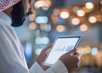 A man in traditional Middle Eastern attire looks at a tablet displaying Fintech 2.0 financial graphs and charts, with a blurred background of warm lights. | FintechZoom