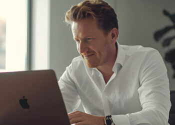 A man in a white shirt smiles while working on a laptop at a desk in a modern, well-lit office, researching if no wagering bonuses could work in traditional casinos, with large windows and a plant in the background. | FintechZoom