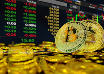 A pile of gold-colored Bitcoin coins in front of a digital stock market screen showing fluctuating numbers, red and green symbols, and colorful financial charts. | FintechZoom