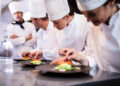 Four chefs in white uniforms and hats are preparing and inspecting dishes on a stainless steel counter in a professional kitchen. Two chefs are plating food, while another observes closely. | FintechZoom