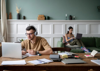 A man wearing headphones works on a laptop at a desk covered with notebooks and papers, while a woman sits on a green sofa in the background using her laptop, highlighting the contrast of working from home vs. office environments. | FintechZoom