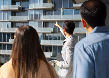Three people, including a man in a light blazer pointing towards upper balconies, stand outside a modern apartment building with glass railings, likely discussing or viewing the property. | FintechZoom