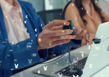 Two people sit at a desk with laptops; one is holding a headset. Currency symbols overlay the image, representing global finance or online transactions. | FintechZoom