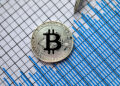 A physical Bitcoin coin placed on top of a financial chart with blue bars and a pen visible, representing cryptocurrency investment and market analysis. | FintechZoom
