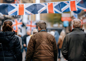 Three people walk down a street decorated with hanging Union Jack flags. The lively scene, with bustling shops and festive crowds, hints at how UK casinos are taking advantage of modern mobile payments during national celebrations. | FintechZoom
