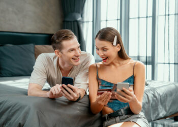 A young man and woman are lying on a bed, smiling and looking excitedly at their smartphones. The woman is holding a few credit cards—perhaps they’re thrilled about an online purchase or discovering how online casinos give out bonuses to players. | FintechZoom
