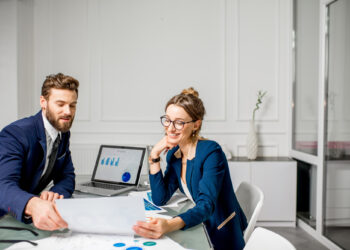 Two business professionals, a man and a woman, sit at a table reviewing papers. A laptop displaying charts is open in front of them. Both are smiling and engaged in discussion in a modern office setting. | FintechZoom
