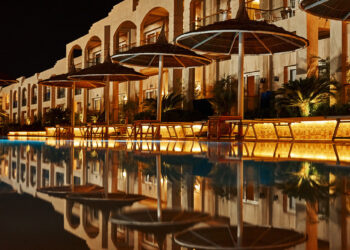 Night view of a lit-up hotel with arched balconies, poolside lounge chairs, and large umbrellas reflecting in a calm swimming pool in the foreground. | FintechZoom