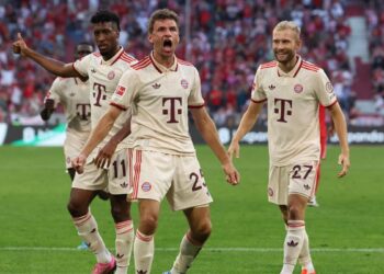 Three Bayern Munich players in white jerseys celebrate on the field after their German Bundesliga win. The central player, Thomas Muller, shouts passionately as the others gesture amid cheers during Bayern Munich's 2-0 victory over SC Freiburg. | FintechZoom