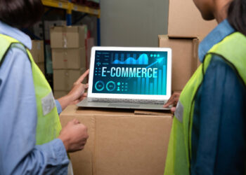 Two people wearing high-visibility vests look at a laptop displaying the word "E-COMMERCE" and data graphics, set on a cardboard box in a warehouse with stacked packages in the background. | FintechZoom