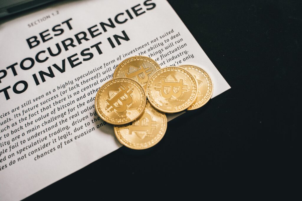 Five gold-colored Bitcoin tokens are placed on top of a printed document titled "Best Cryptocurrencies to Invest In." The coins partially cover the text, and the background is dark. | FintechZoom