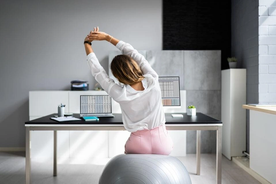 A woman in a white blouse and pink pants practices chair yoga, stretching her arms overhead while sitting on an exercise ball at her desk, facing two computer monitors displaying spreadsheets. | FintechZoom