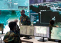 Soldiers in camouflage uniforms, alongside defense contractors, monitor multiple computer screens displaying maps and aerial surveillance data in a modern military command center with large digital displays on the walls. | FintechZoom