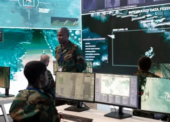 Soldiers in camouflage uniforms, alongside defense contractors, monitor multiple computer screens displaying maps and aerial surveillance data in a modern military command center with large digital displays on the walls. | FintechZoom