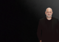 Bruce Buchanan, a bald man with a short beard, wears a black suit and shirt as he stands against a gradient black-to-gray background, positioned on the right side of the image with his hands in his pockets. | FintechZoom