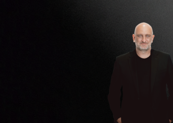 Bruce Buchanan, a bald man with a short beard, wears a black suit and shirt as he stands against a gradient black-to-gray background, positioned on the right side of the image with his hands in his pockets. | FintechZoom