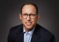 Glenn Lurie, wearing glasses and a dark pinstripe suit with a light blue shirt, smiles at the camera against a plain dark background. | FintechZoom