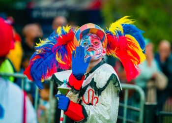 A person in colorful clown makeup and costume, with large red, blue, and yellow feathers on their hat, raises a blue-gloved hand while holding a decorated staff at an outdoor event celebrating one of the Best Cultural Festivals to Celebrate in Africa. | FintechZoom