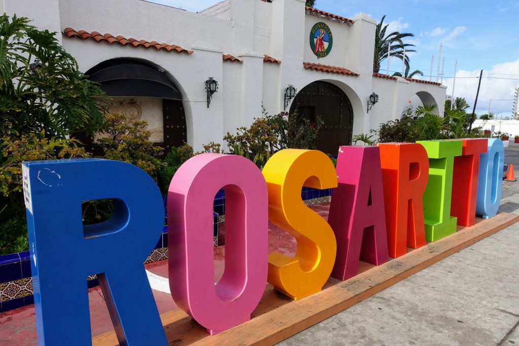 Large, colorful letters spelling "ROSARITO" stand before a white building with a tiled roof, lush plants, and a decorative emblem—perfect for capturing photos after exploring the region’s renowned wine country under the partially cloudy sky. | FintechZoom