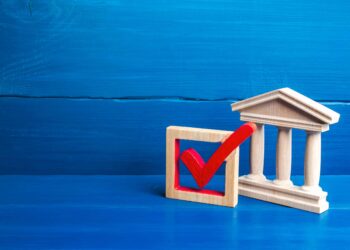 A wooden model of a government building represents Neobanks vs Traditional Banks, placed next to a wooden square featuring a bold red checkmark, set against a blue wooden background. | FintechZoom