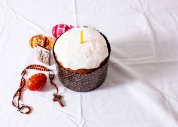 A decorated Easter cake with white icing and a single yellow candle is placed on a white cloth, surrounded by painted Easter eggs, a small religious icon, and a wooden rosary with a cross. | FintechZoom