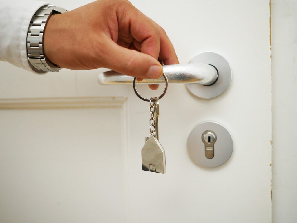 A hand holding a keyring with a house-shaped keychain, inserting a key into a door lock—an image that captures the essence of real estate management. The door and lock are white and silver; the person wears a white shirt and silver watch. | FintechZoom