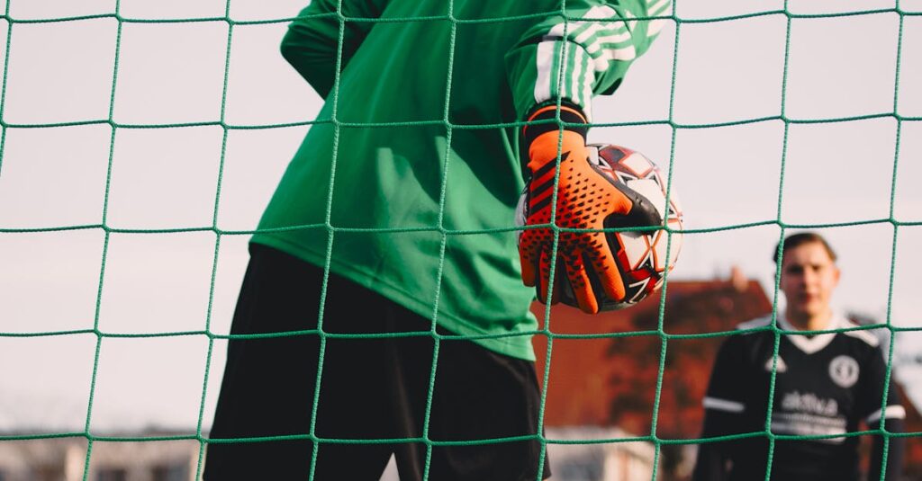 A soccer goalkeeper wearing an orange glove and green jersey holds a ball behind a goal net, with another player in a black uniform standing in the background. | FintechZoom