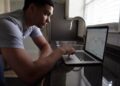 A man leans on a kitchen counter, focused on a laptop displaying a candlestick stock chart next to a sink and window with blinds. | FintechZoom