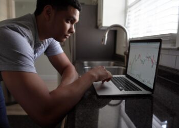A man leans on a kitchen counter, focused on a laptop displaying a candlestick stock chart next to a sink and window with blinds. | FintechZoom