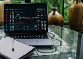 A laptop displaying cryptocurrency trading charts and data sits on a glass table next to a notebook with a pen, subtly hinting at both crypto gambling and trading, with decorative vases and greenery visible in the background. | FintechZoom