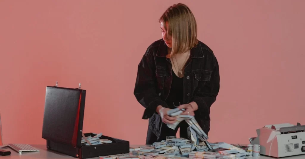 A woman in dark clothing stands at a table covered with stacks of cash, placing some bundles into an open black briefcase against a pink background. | FintechZoom