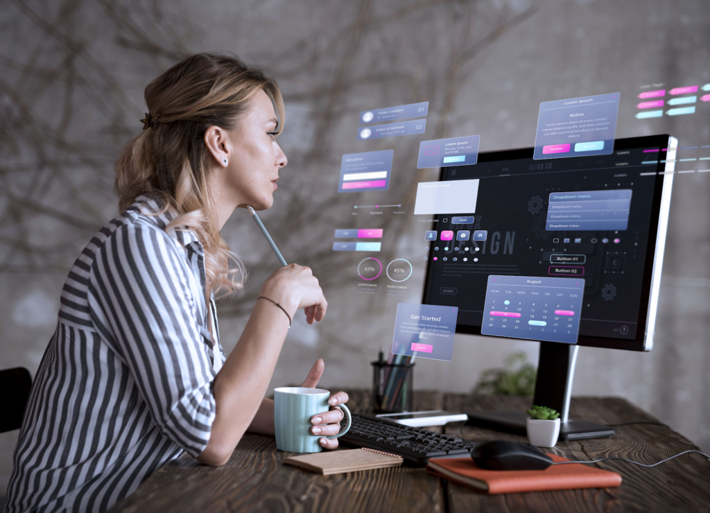 A woman sits at a desk using a desktop computer, interacting with floating digital interface windows. She holds a pencil and mug, focusing on Transcription tasks on the futuristic screen in a modern office setting. | FintechZoom