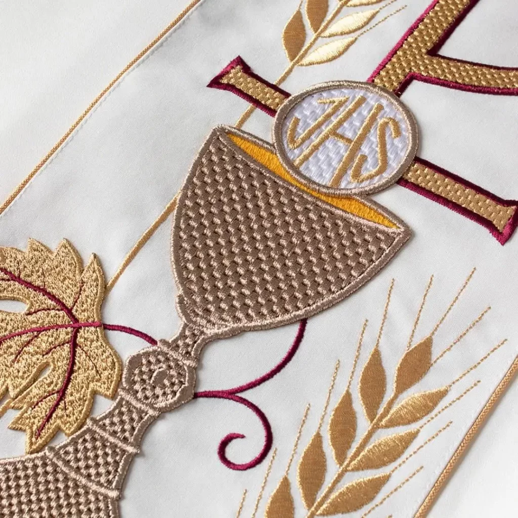 Detailed embroidery of a chalice, wheat, grapes, and a cross with "IHS" on white fabric, symbolizing Christian Eucharist. Gold, brown, and maroon threads enrich this classic motif often seen on liturgical vestments. | FintechZoom