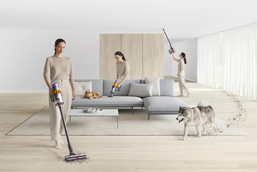 A woman is shown multiple times vacuuming a modern living room with a cordless vacuum cleaner, cleaning up after a dog and cat on light wood floors around a gray couch. | FintechZoom
