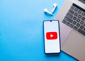 A smartphone displaying the YouTube logo is placed next to a laptop and a pair of wireless earbuds on a blue surface. | FintechZoom