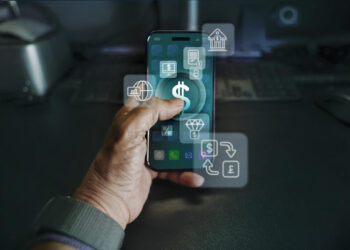 A hand holds a smartphone displaying financial icons, including a large dollar sign, while Fintech Tools and digital banking symbols float around the screen. A keyboard and computer setup are visible in the background. | FintechZoom
