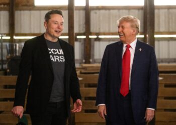 Two men stand indoors, smiling and conversing—reminiscent of a casual meeting between Musk and Trump. One wears a dark jacket and graphic T-shirt, holding a cap; the other sports a blue suit with red tie. Wooden benches and metal structure are behind them. | FintechZoom