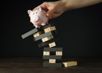 A hand places a pink piggy bank on top of a precarious, wobbly stack of wooden blocks, illustrating financing challenges and financial instability against a dark background. | FintechZoom