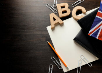 Wooden letters A, B, and C, a pencil, paper, black notebooks, paper clips, and a small UK flag are arranged on a dark wooden desk—perfect for discussing English Proficiency Tests. The left side of the image is mostly empty for copy space. | FintechZoom