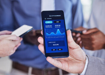 A person holds a smartphone displaying a cryptocurrency trading app with Bitcoin and USD values and a price chart, while two others nearby use their phones. | FintechZoom