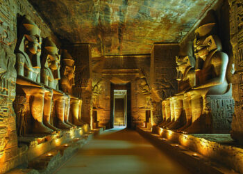 Ancient Egyptian temple interior with large seated statues lining both sides, illuminated by warm golden light. The walls and ceiling are covered with detailed hieroglyphics and carvings. | FintechZoom