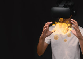 A person wearing a virtual reality headset and a white t-shirt interacts with floating digital blocks and golden Bitcoin symbols on a dark background. | FintechZoom