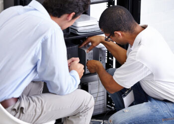 Two men, resembling ATM maintenance providers, are kneeling and working on computer servers in a server rack. One points at equipment while the other observes closely, both focused on precise technical maintenance. | FintechZoom