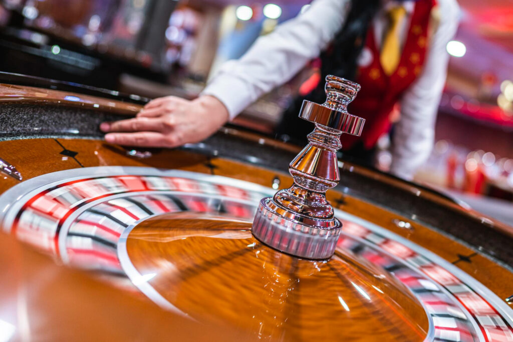 A close-up of a spinning roulette wheel in a casino, with a croupier in uniform reaching out to stop the wheel. The colorful numbers blur from the motion, creating a dynamic and vibrant atmosphere. | FintechZoom